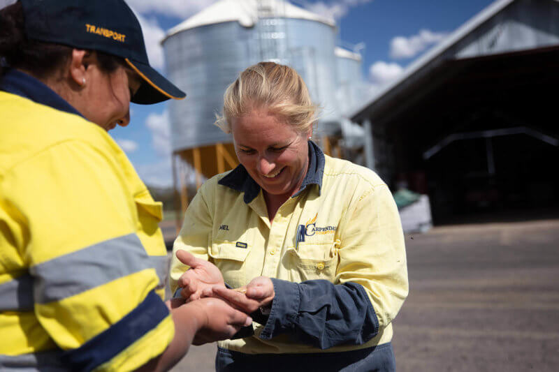 Two women wear high-vis clothing looking down at their hands cupped holding grain on farm with silo and shed in the background.